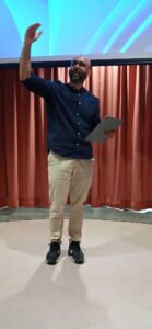Man in a blue shirt and khakis gestures while holding a tablet at a presentation.